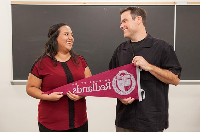 Two people smile at each other in a classroom while holding a pennant flag from the University of Redlands.